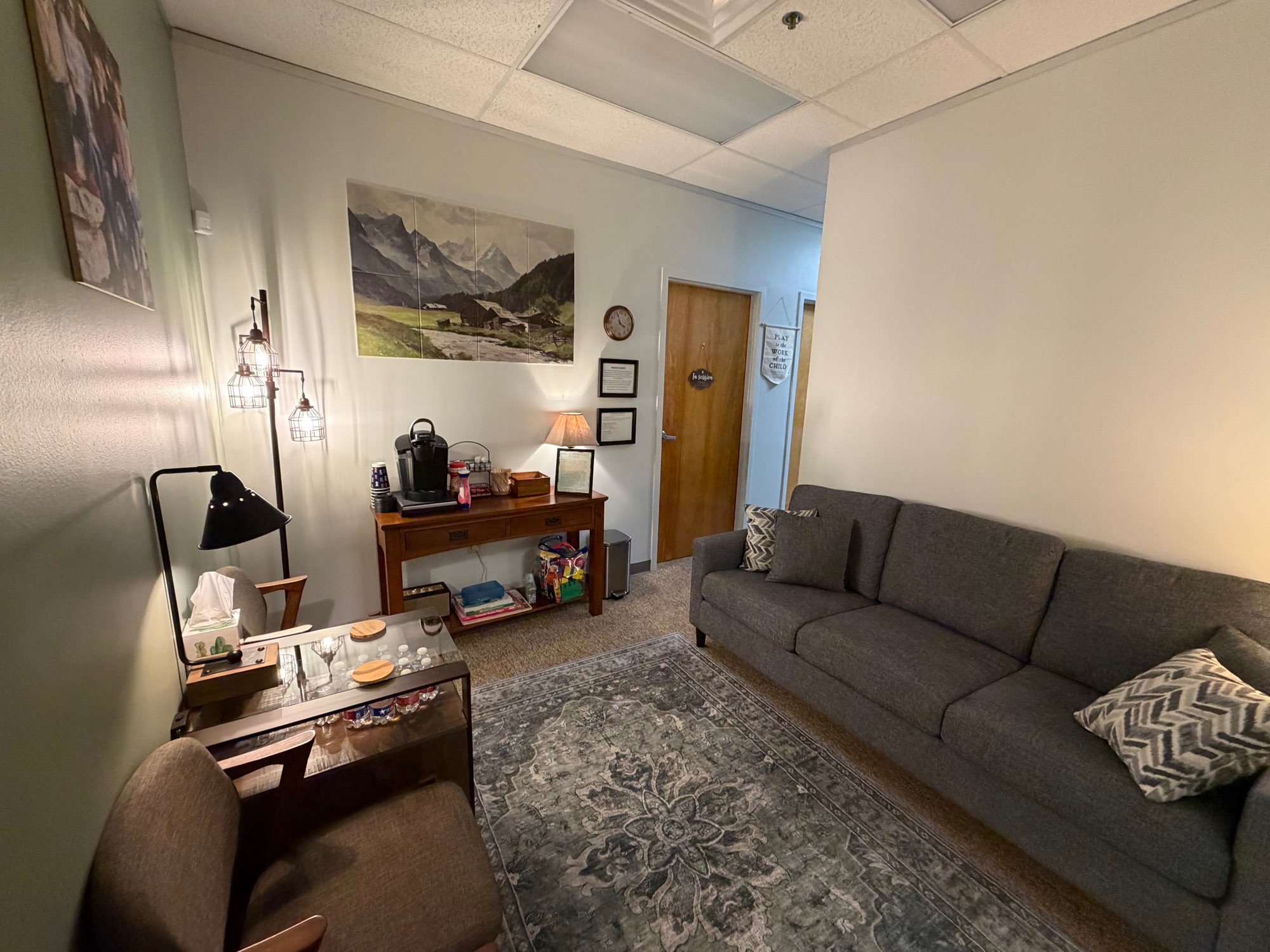 Waiting room with a gray couch, patterned rug, coffee station, and a mountain landscape on the wall